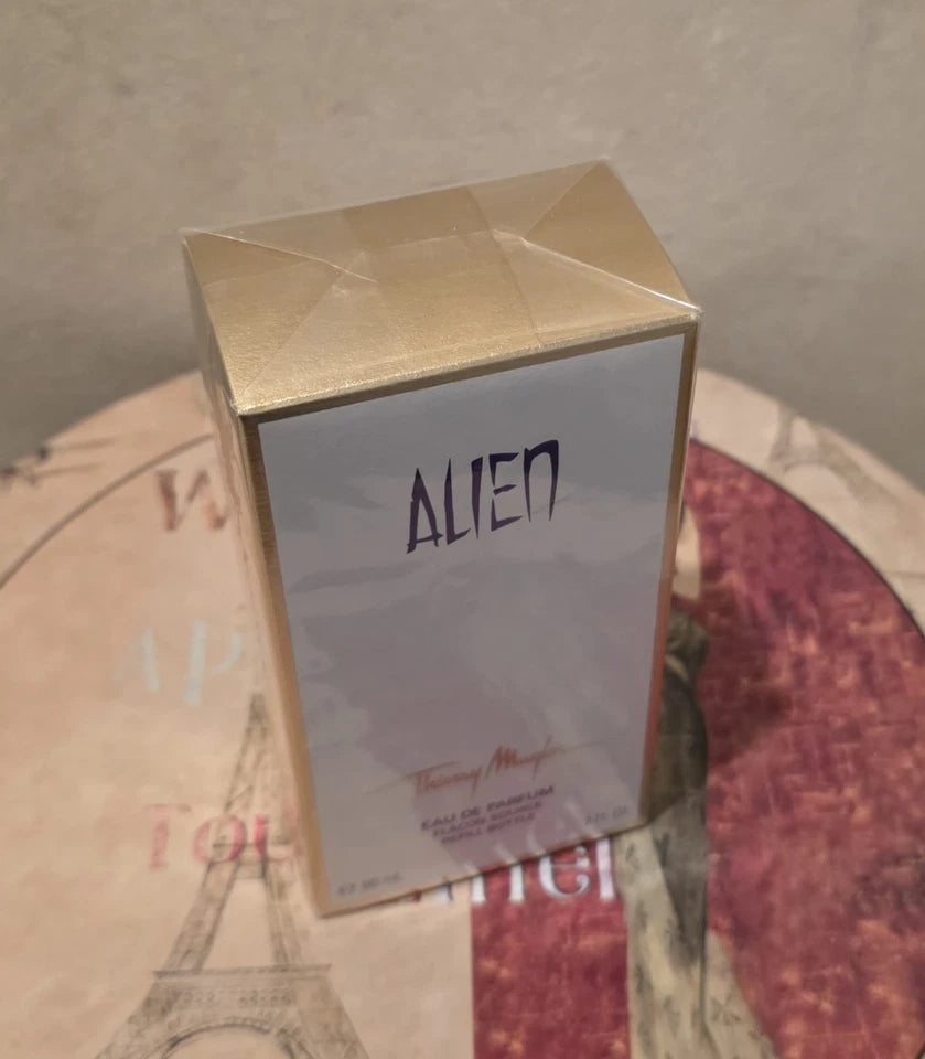 A boxed perfume product labeled Alien, with a metallic gold top and a pale lavender front panel, photographed on a patterned round surface against a neutral wall.