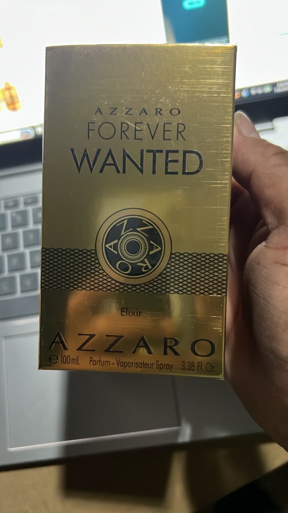 A hand holding a gold Azzaro Forever Wanted Elixir perfume box in front of a laptop and desk background.